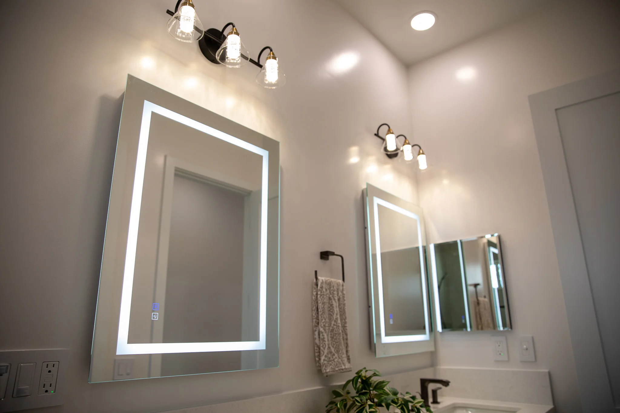 Dual LED-backlit bathroom mirrors with black sconce lighting and recessed ceiling lights